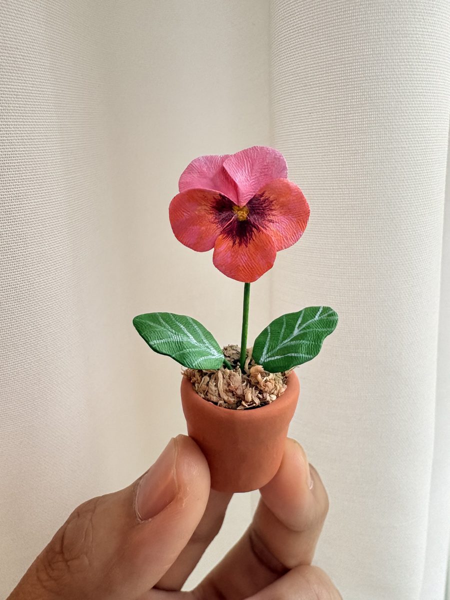 A tiny clay pink pansy in a thimble-sized terracotta pot, held between two fingers.