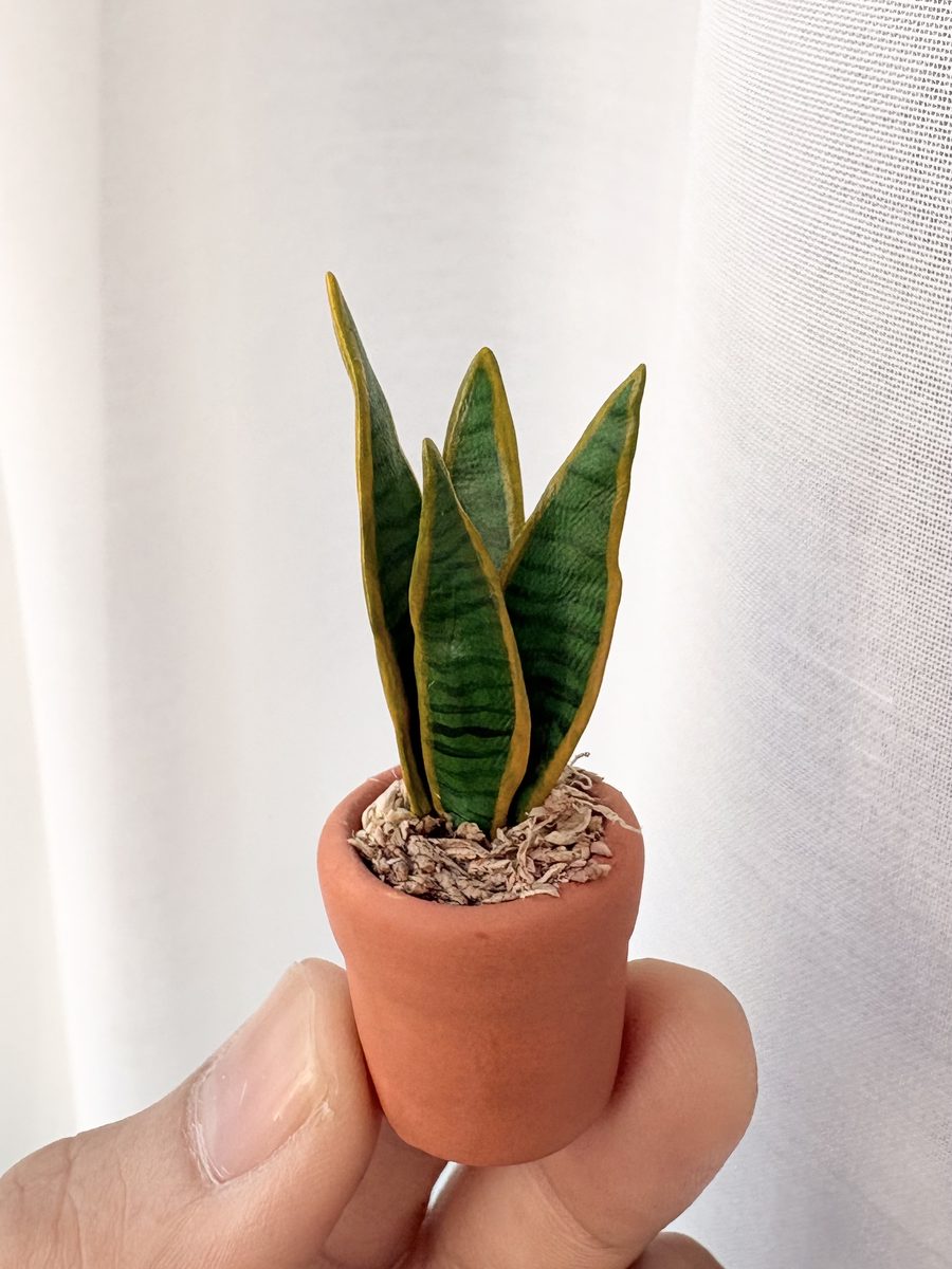 A tiny clay snake plant with striped upright leaves, held between two fingers.